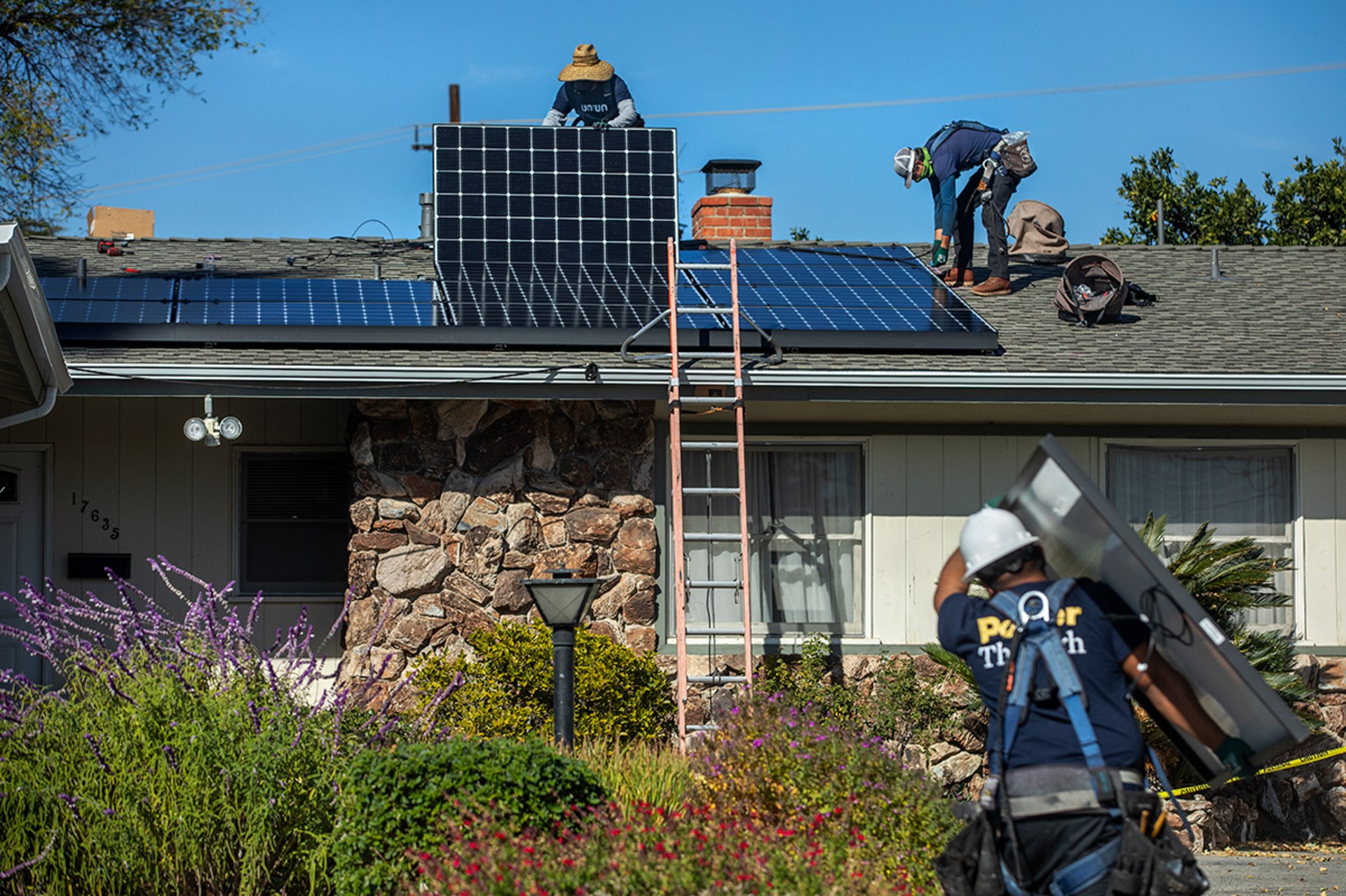 Two workers install solar panels on the roof of a house while one worker walks towards them carrying another solar panel.