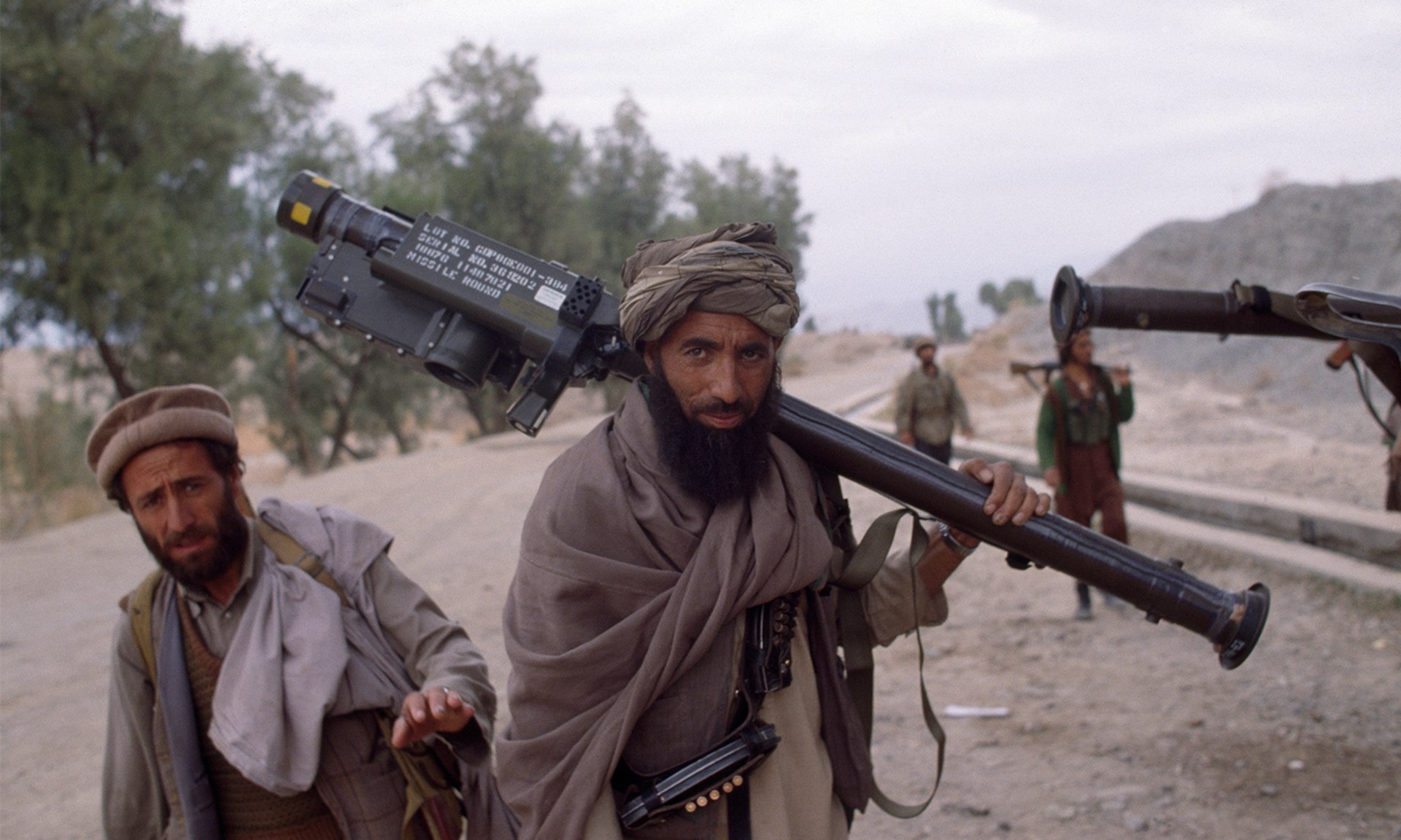 Bearded man in cloak and turban with several other men walks down dusty road while carrying stinger missile launcher over his shoulder.