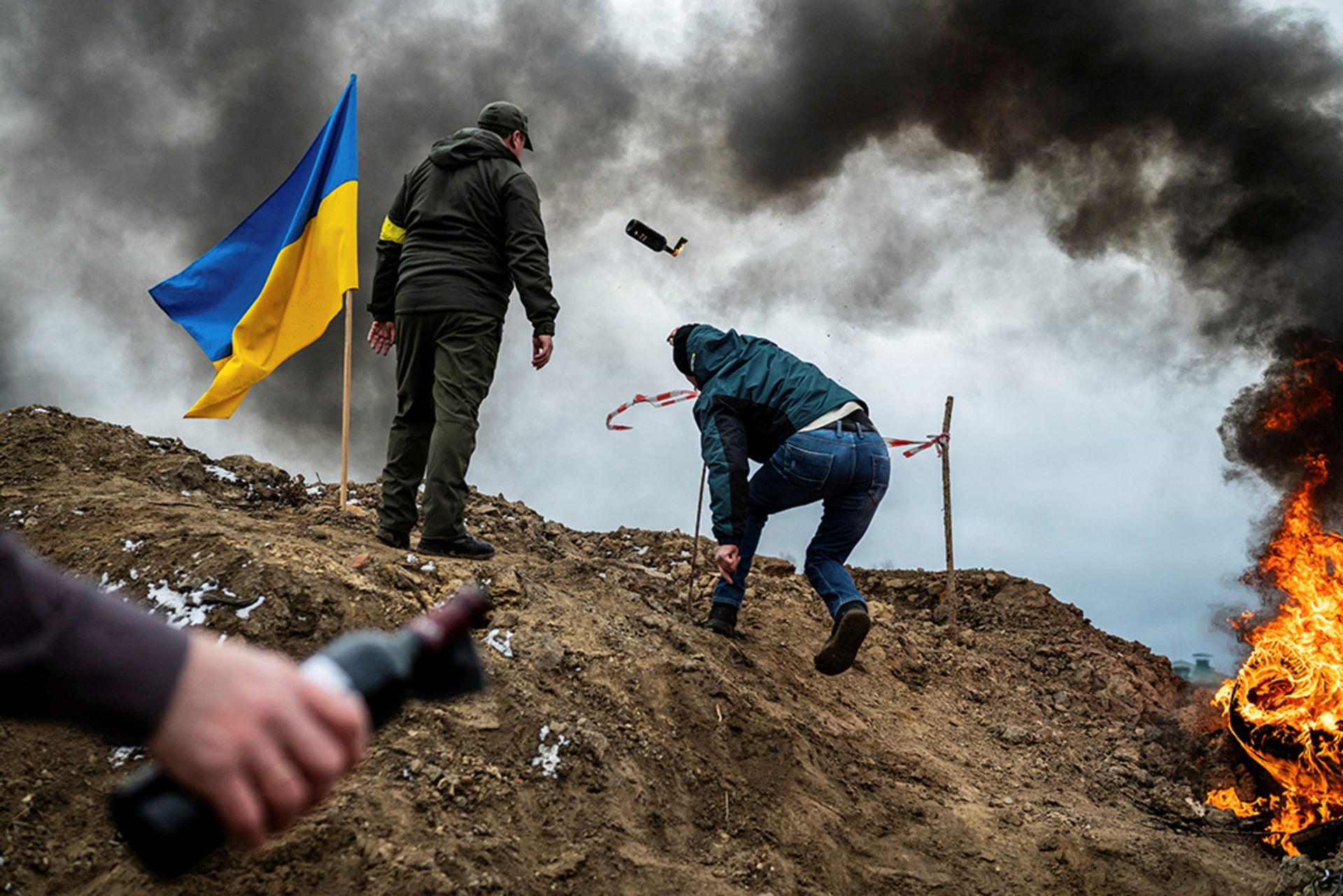 Two soldiers in smoke on battlefield next to Ukrainian flag. One of them throws a Molotov cocktail.