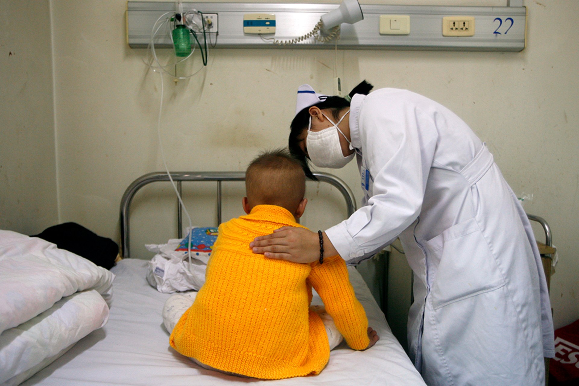 A nurse comforts a young patient with her hand on their back as they sit on a bed.