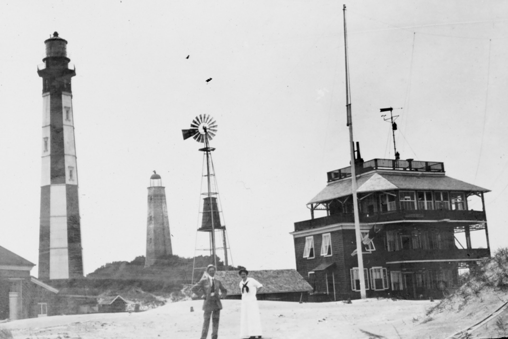Two people stand in front of the weather station at Cape Henry, Virginia circa 1900, which consists of a building, lighthouses, and a weather vane.