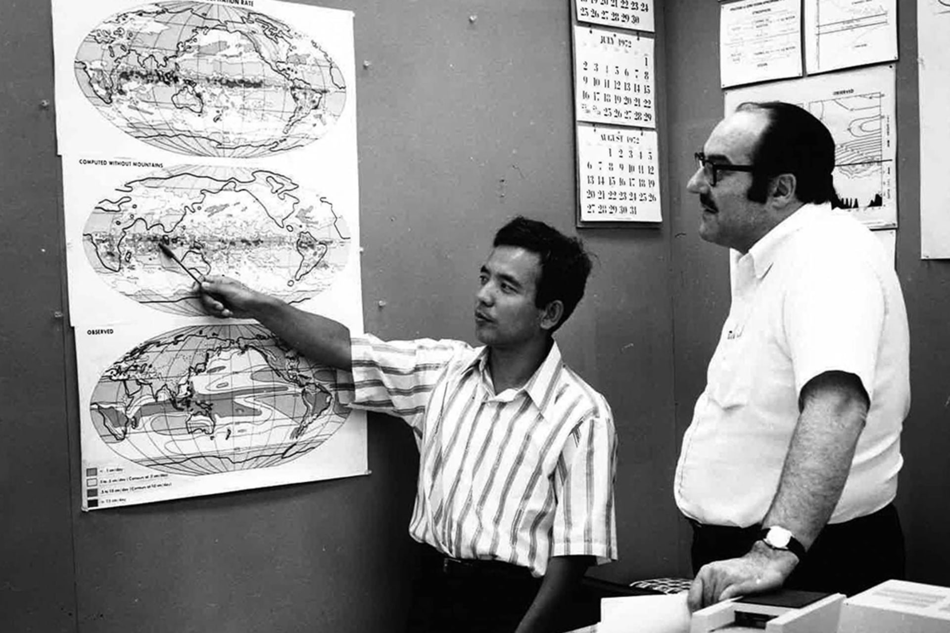 Syukuro Manabe, a scientist, points to a map that models climate change as another man stands next to him looking at the map.