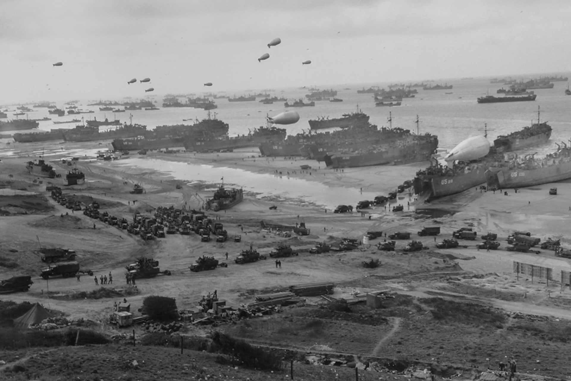 U.S. landing ships bring cargo ashore as barrage balloons float overhead to protect the ships and trucks head inland on one of the invasion beaches in Normandy in June 1944.