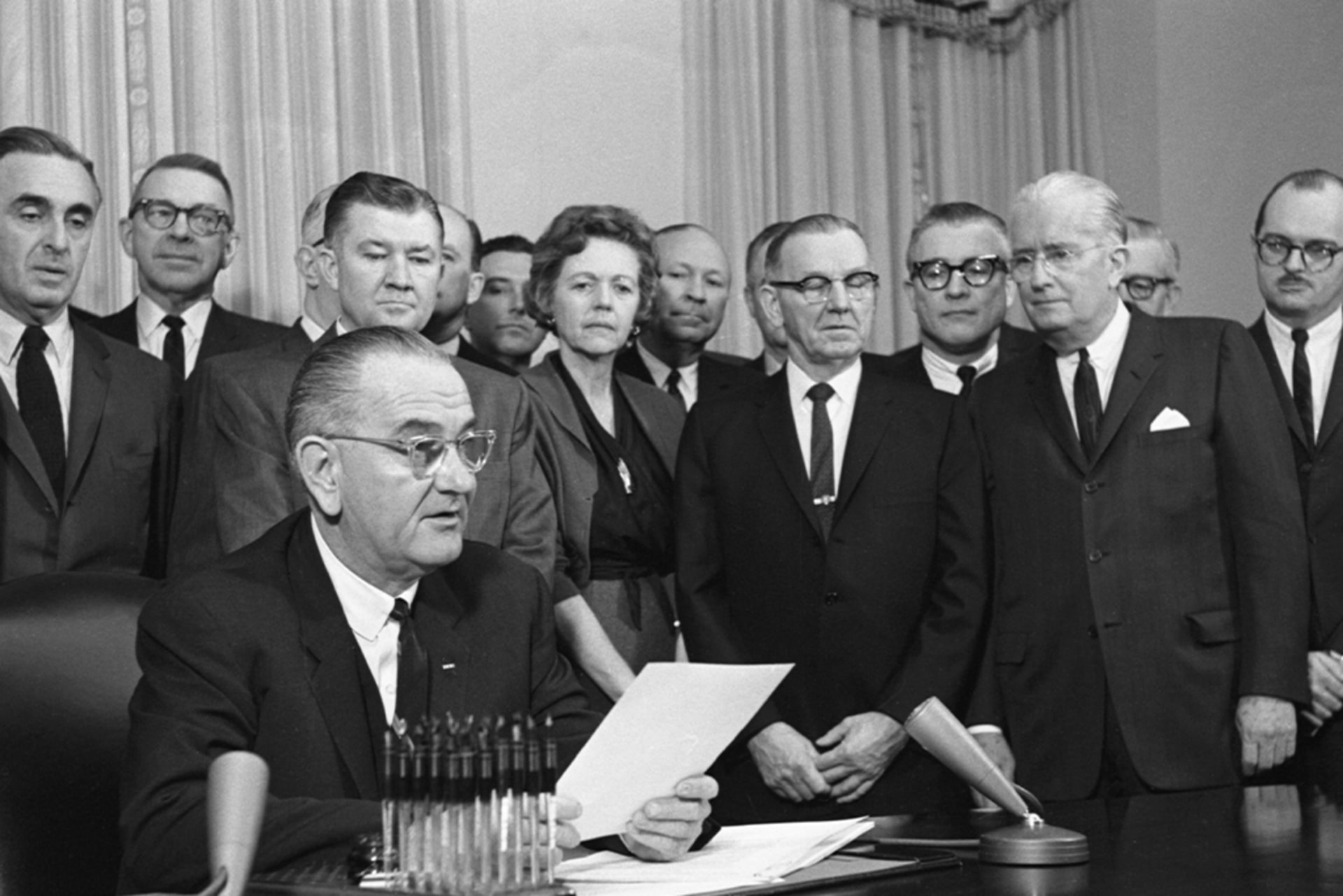 President Lyndon Johnson signs the Clean Air Act into law on December 17, 1963, as various people stand behind him and look on.