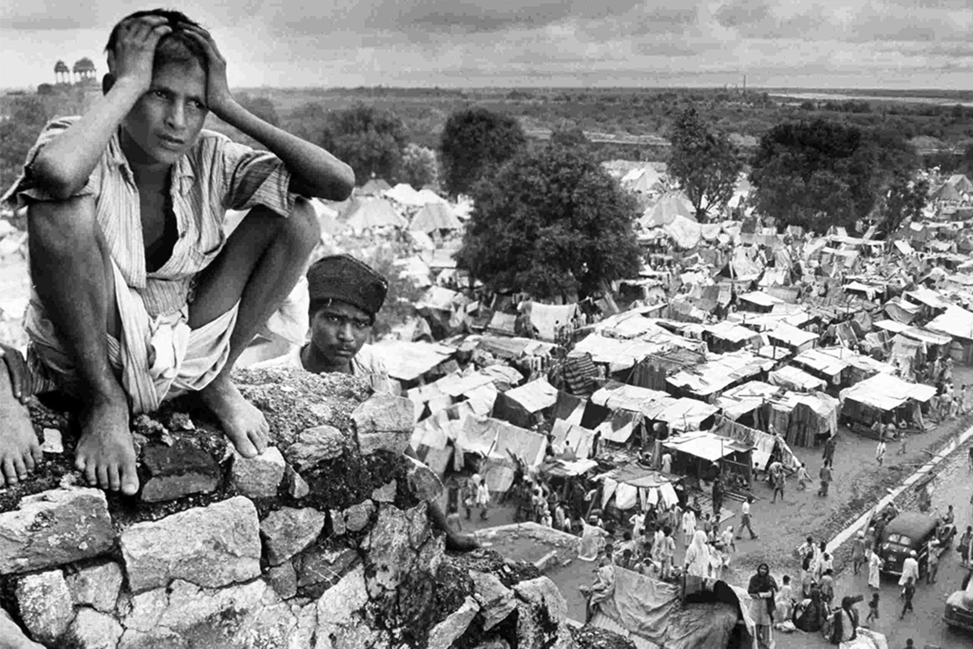A distraught boy sat on the walls of a refugee camp in Delhi during the Partition of India in 1947. The refugee camp can be seen behind and to the right of him. Another boy peeks over the wall.