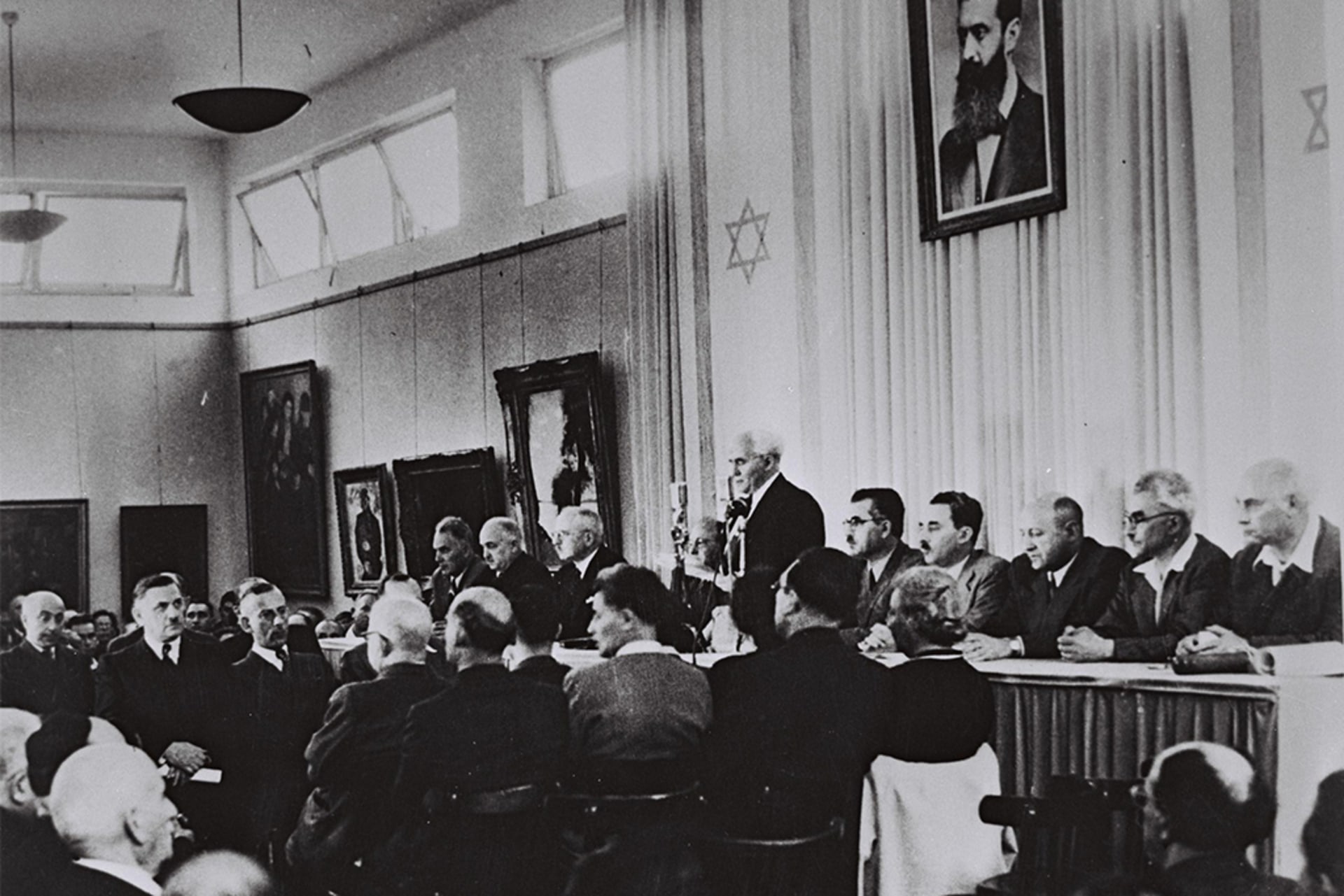 David Ben-Gurion, flanked by the members of his provisional government, reads the Declaration of Independence in the Tel Aviv Museum Hall. The flag of Israel can be seen hanging on the wall behind him.