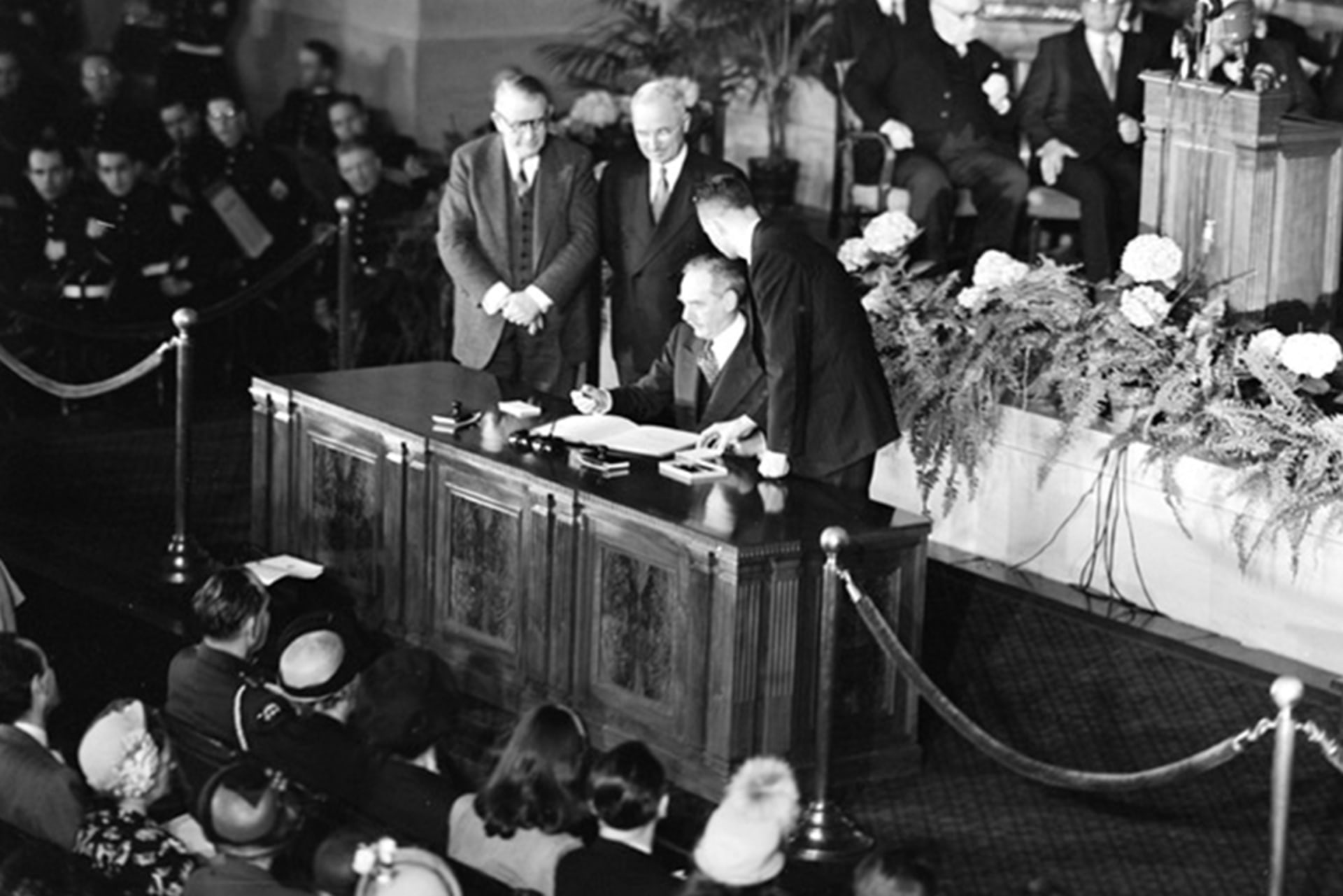 Secretary of State Dean Acheson signs a treaty, President Harry Truman and Vice President Alben Barkley are standing next to him.