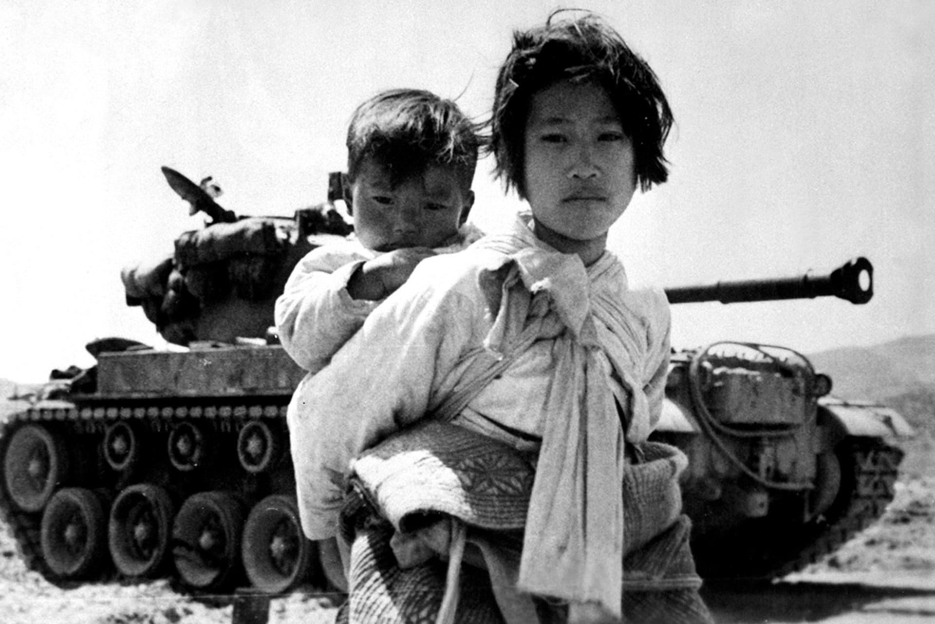 Black and white photo of a young Korean girl holding a toddler on her back, while standing in front of a tank.