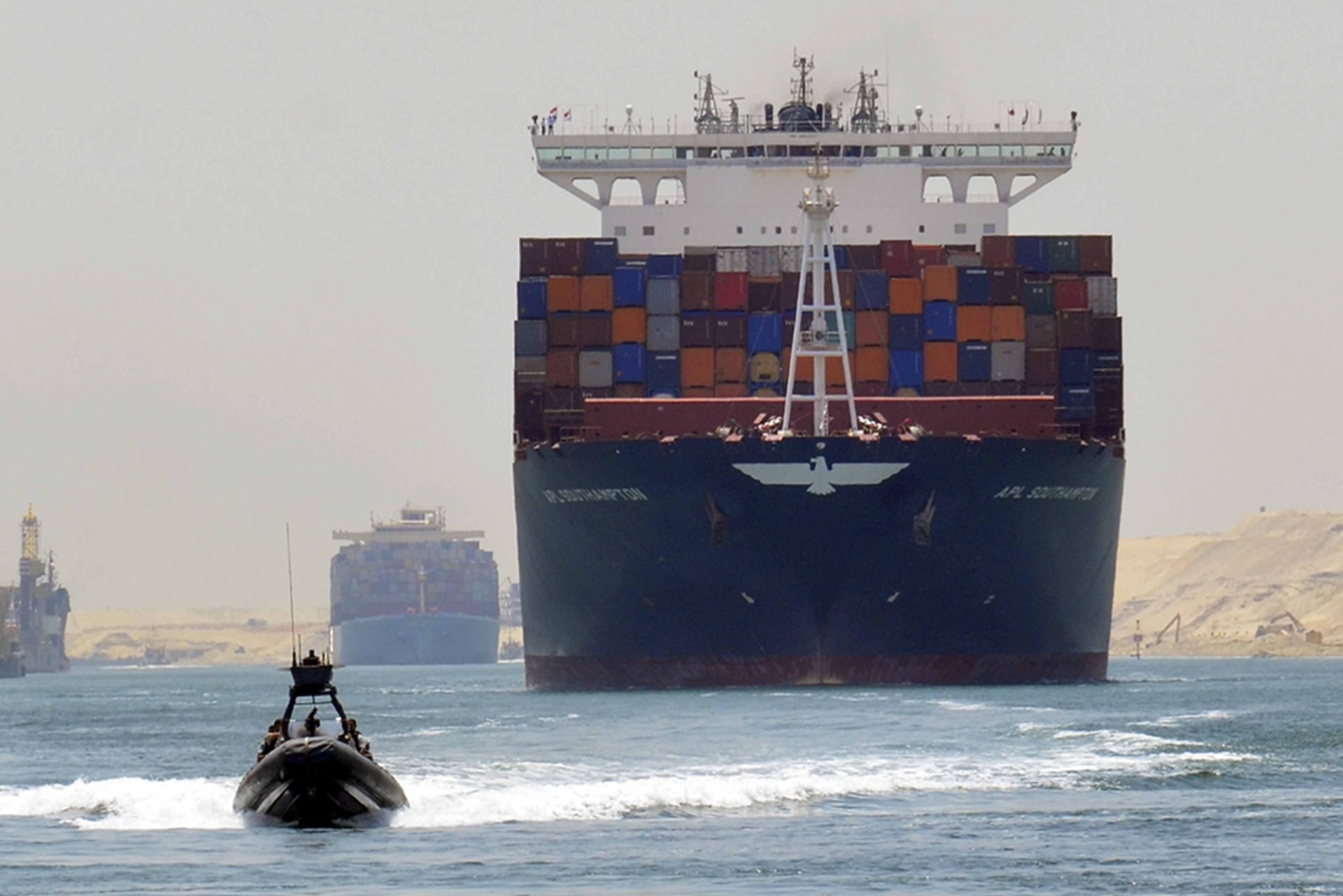 A cargo ship is seen crossing through the Suez Canal. A smaller motor boat drives in front of it and another cargo ship trails behind it.