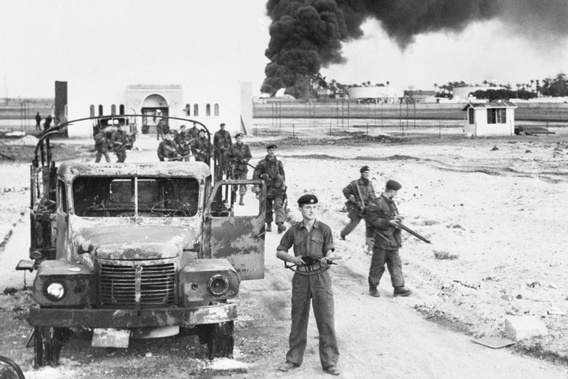 A photo of British troops invading Egypt and oil installations burning at Port Said, north of the Suez Canal. The men hold guns and survey their surroundings; there is a large plume of smoke erupting from a burning building in the back.