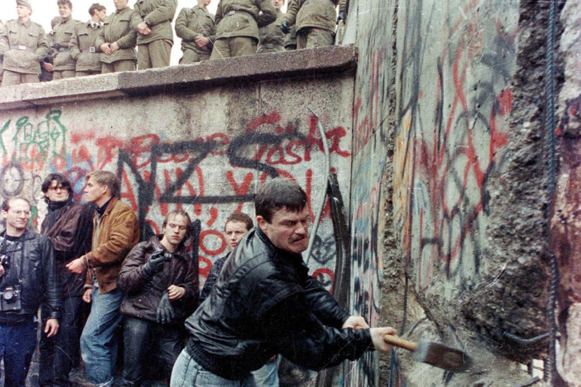 A demonstrator pounds away the Berlin Wall as East Berlin border guards look on from above the Brandeburg Gate.