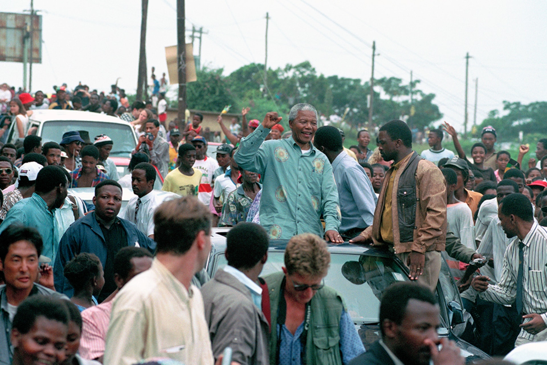 Nelson Mandela sits in the sunroof of a car and raises his fist to acknowledge a crowd.
