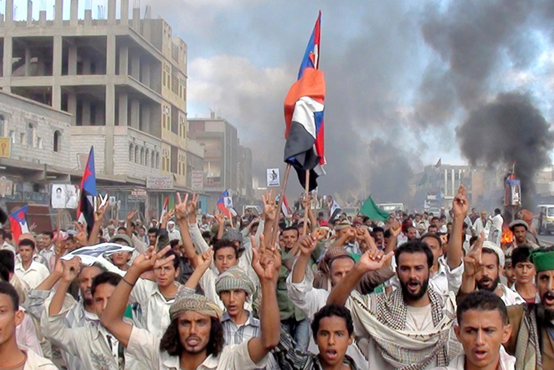 Protestors hold up their hands, and one holds a Yemeni flag, during an anti-government demonstration. There are plumes of smoke coming out of various places behind them.