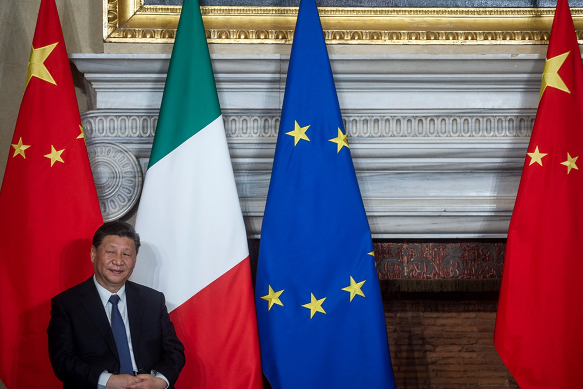 Chinese President Xi Jinping sits and poses in front of Chinese, Italian, and European Union flags.