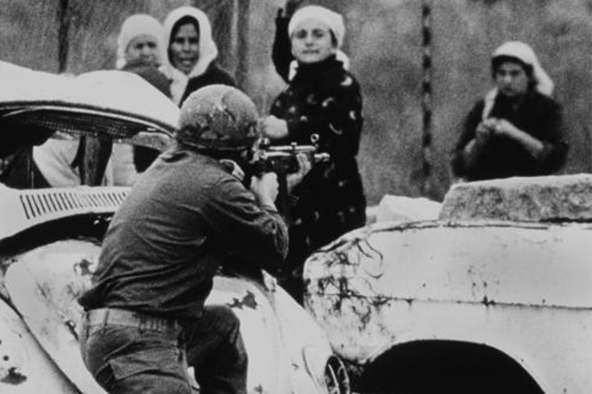 An Israeli soldier ducked behind a broken down car and takes aim at a Palestinian woman standing in a group. The woman stands poised to hurl a rock at him.