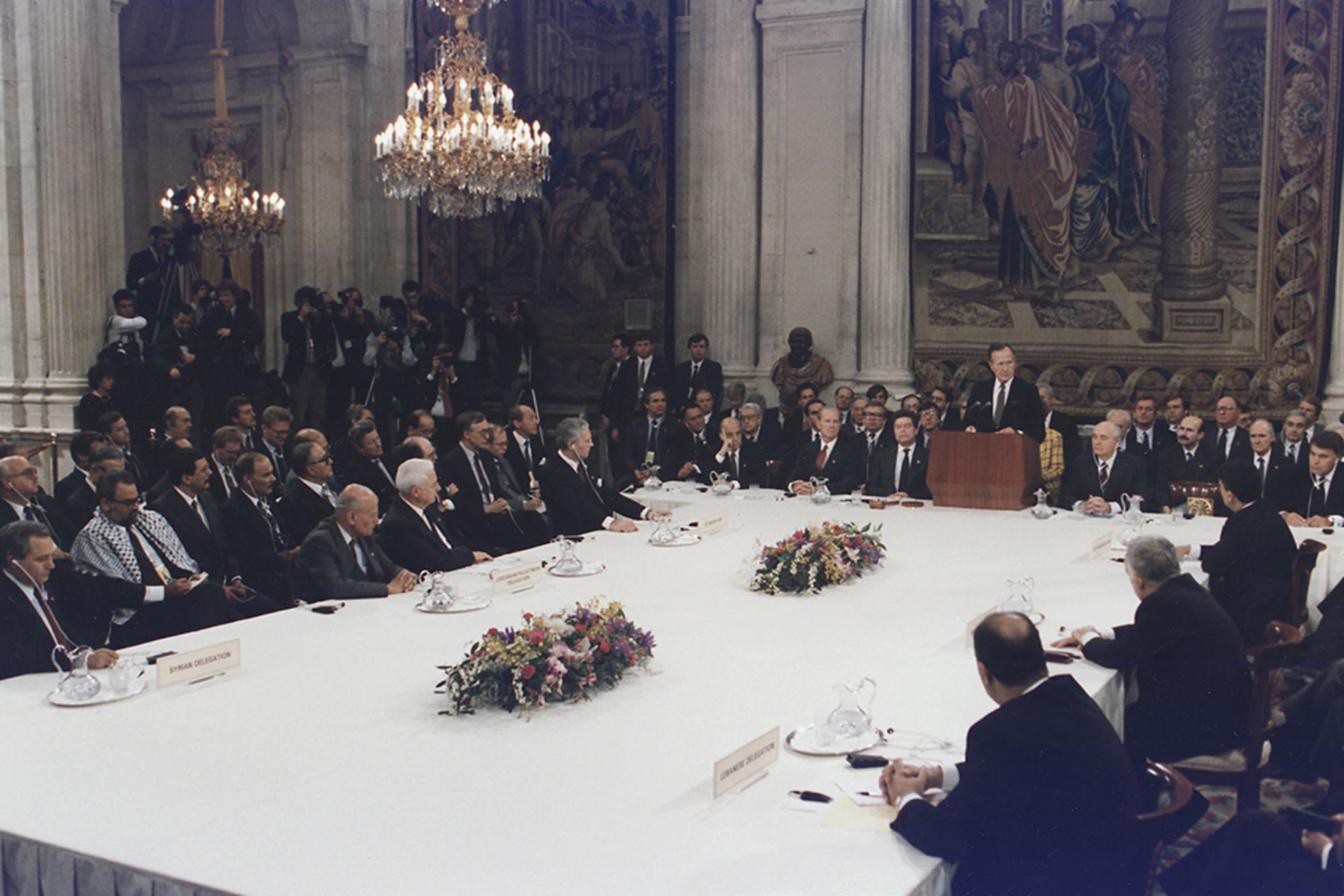 President Bush addresses the Middle East Peace Conference at the Royal Palace. The group sits in a grand room around a large white table beneath chandeliers and tapestries.