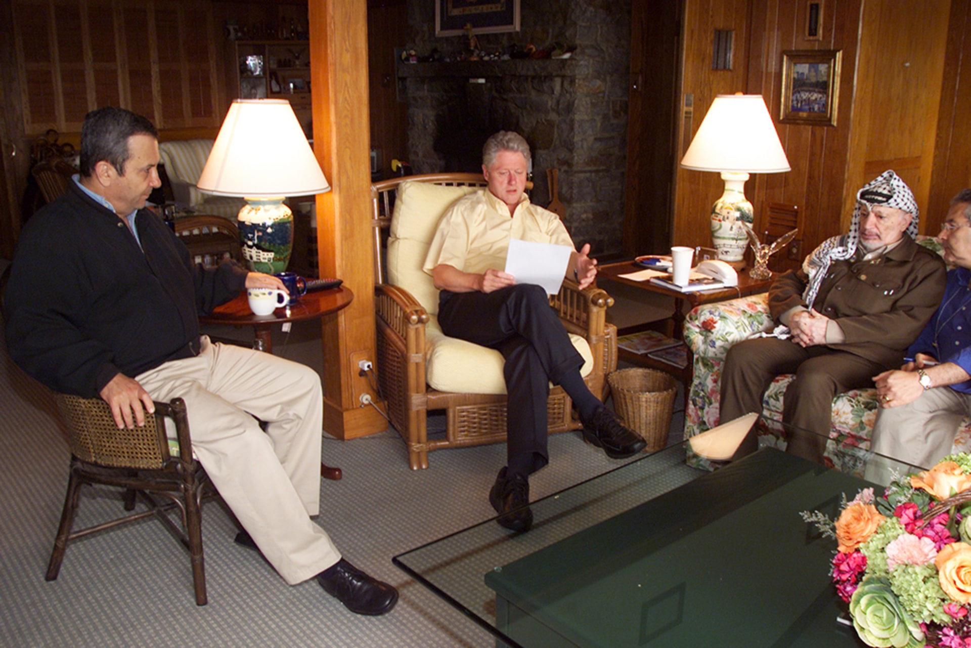 President Clinton meets with Israeli Prime Minister Barak and Palestinian President Yasser Arafat. The leaders sit together in a living room. Clinton reads a document aloud while his fellow leaders listen on.