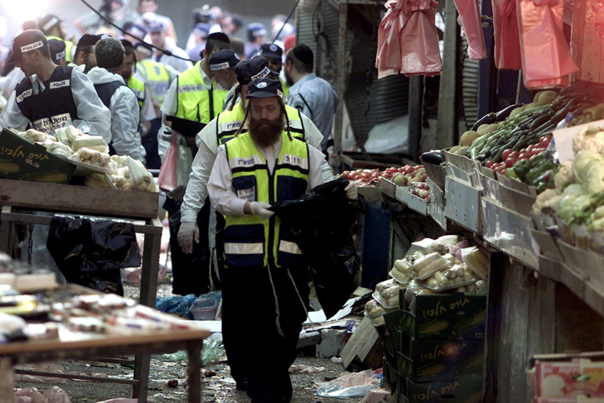 Israeli rescue workers search the scene of a suicide bombing in Netanya. The workers walk amongst the destruction at a vegetable market; produce and miscellaneous debris lays scattered at their feet.
