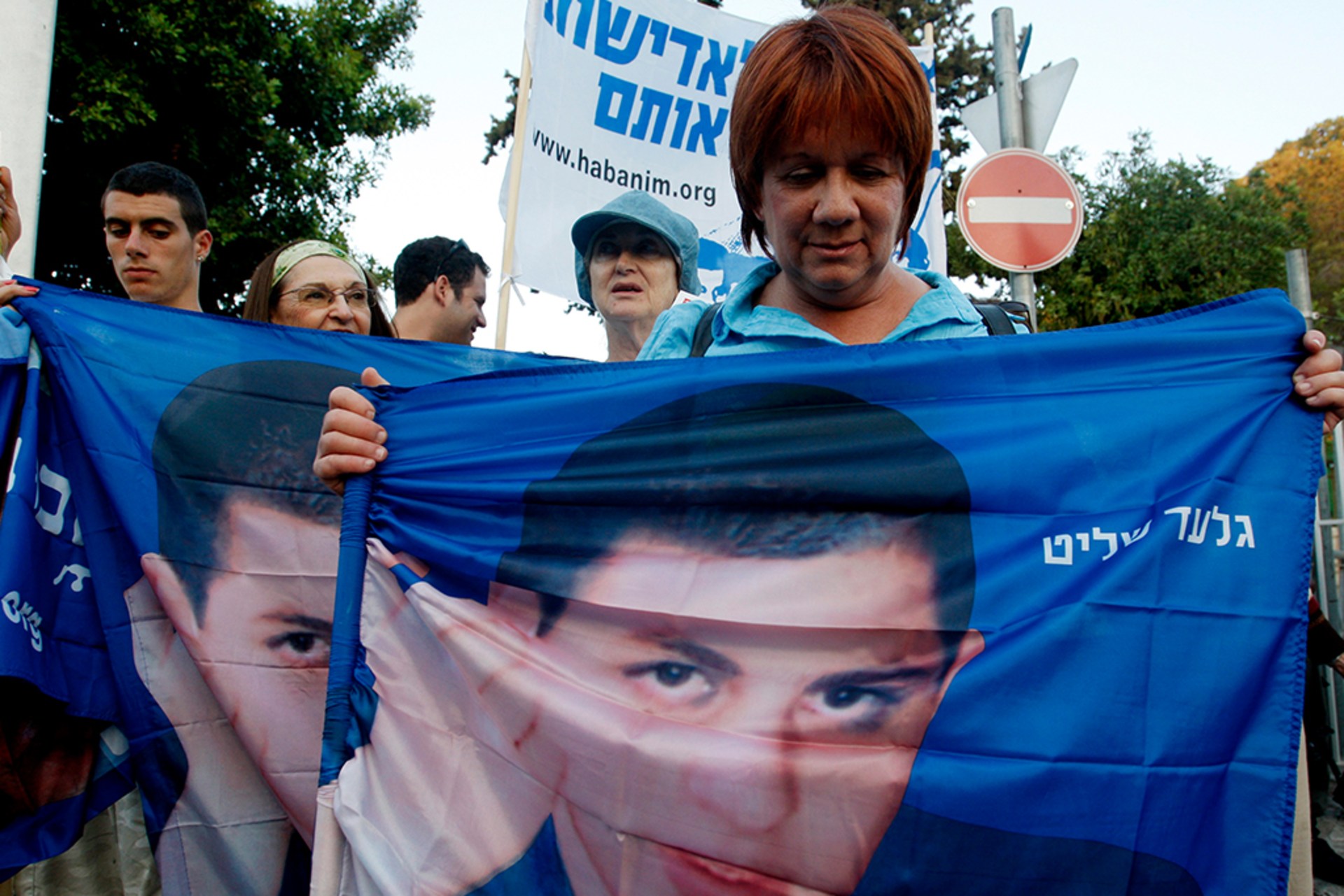 People attend a rally marking two years since Israeli soldier Gilad Shalit was captured by Palestinian militants on the Israel-Gaza border. A woman holds out her flag, displaying Shalit's face. Fellow protestors hold similar banners in the background.