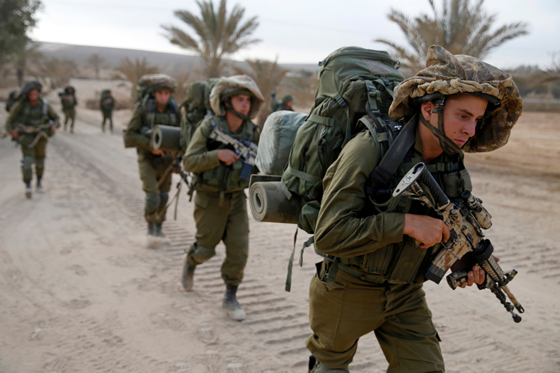 Israeli soldiers walk outside the Gaza Strip as they proceed towards Gaza. The soldier leading has a serious expression and looks down. The soldiers wear camouflage with their weapons held at the ready.