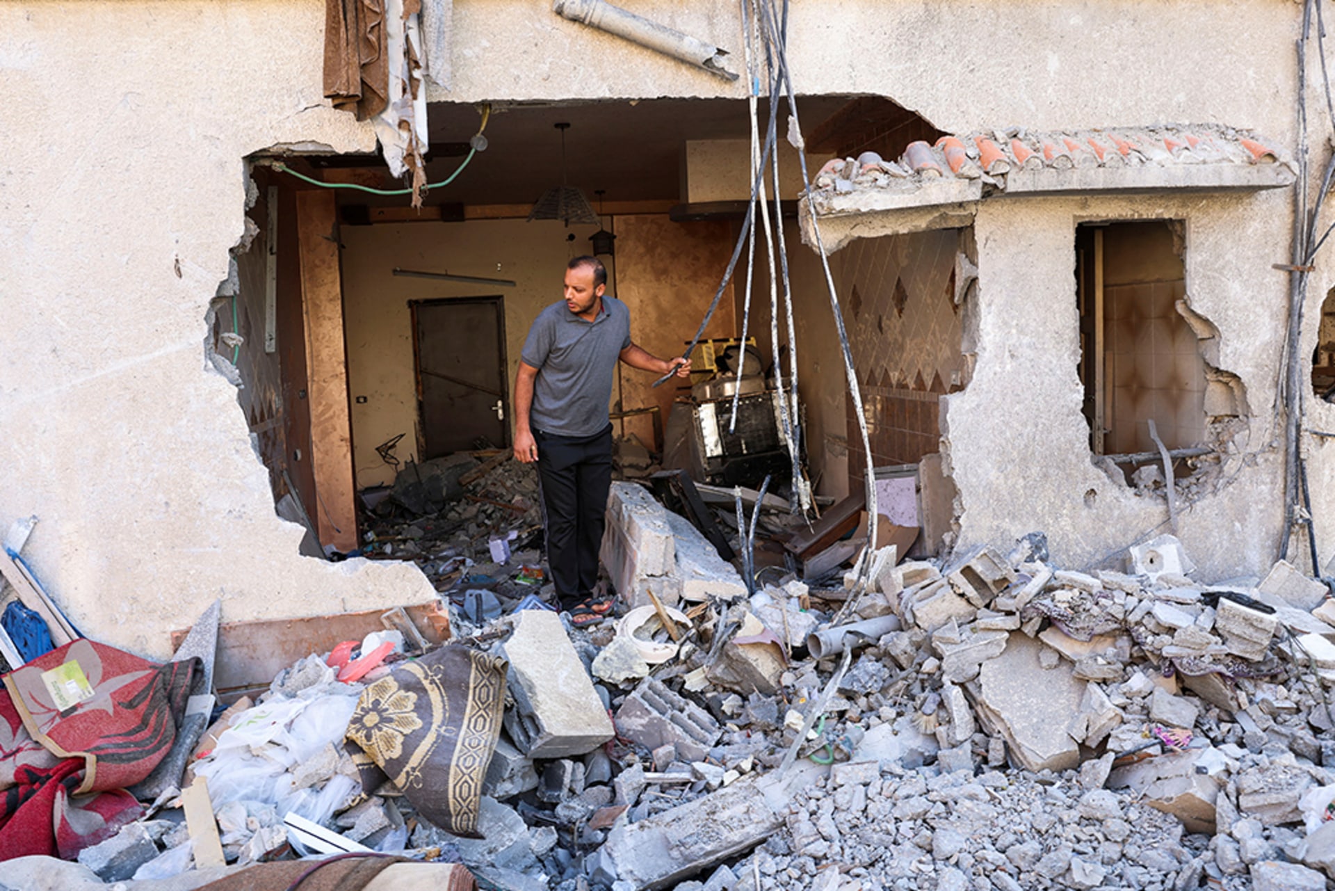 A Palestinian man checks a house that was damaged during Israel-Gaza fighting. The man peers out of a large hole in the side of the house. At his feet, there is a large pile of rubble and scattered debris from within the house.