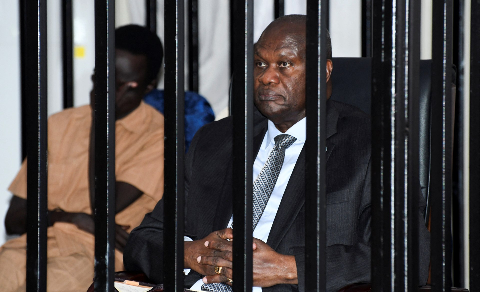 A man wearing a suit sits inside a black steel cage.
