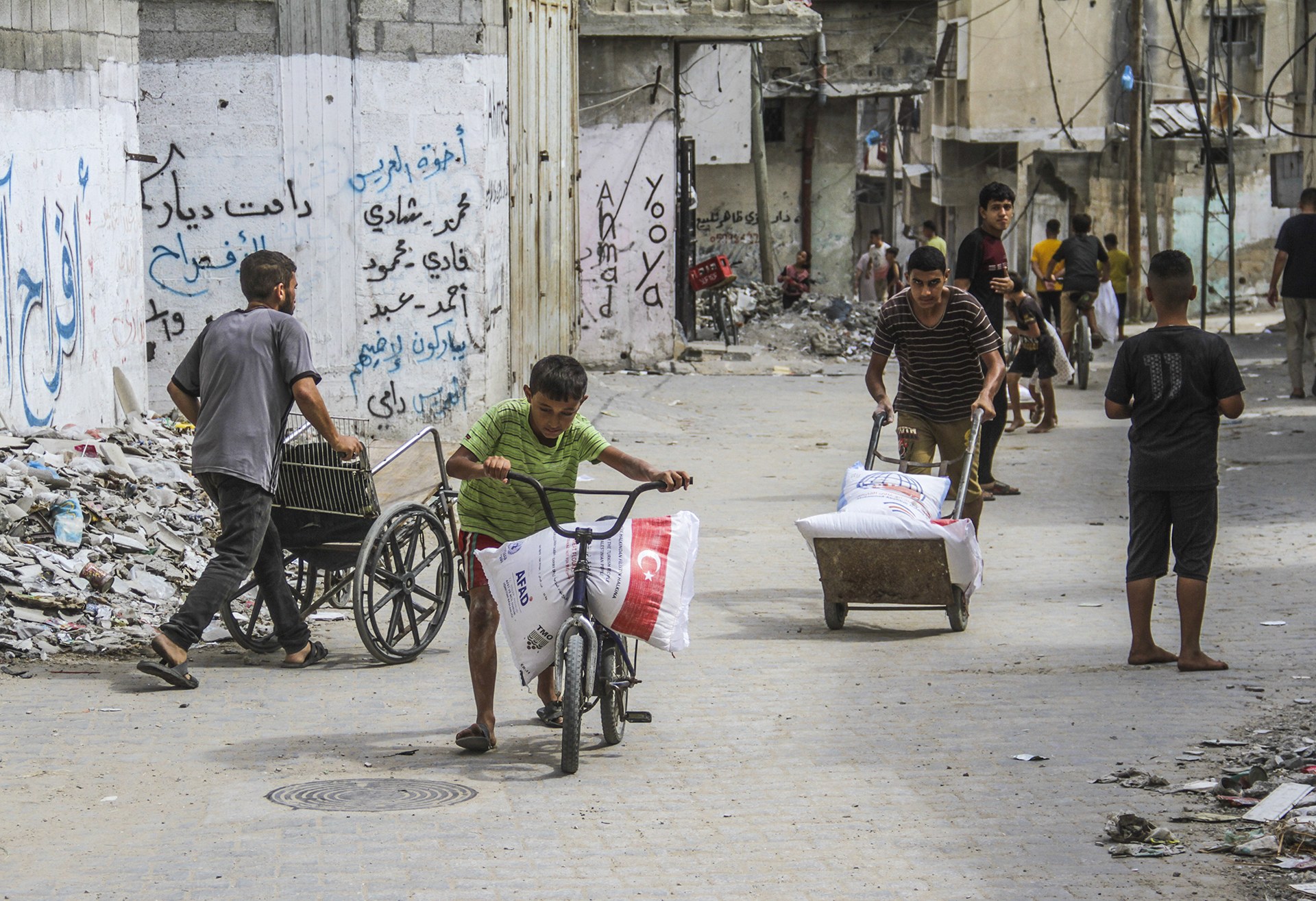 A young boy wheels a bike carrying a bag of food aid along a street, with others following him back to their camp.