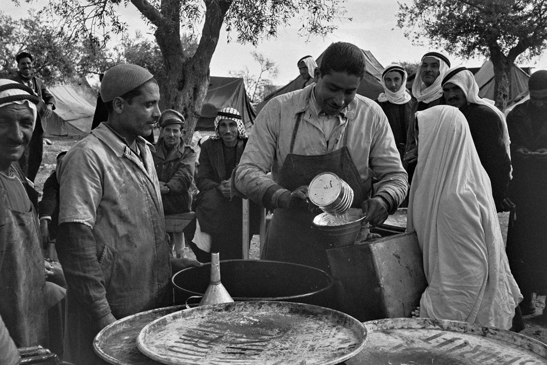 A black and white photo shows a man providing supplies, like food and water, at a distribution point in a refugee camp.