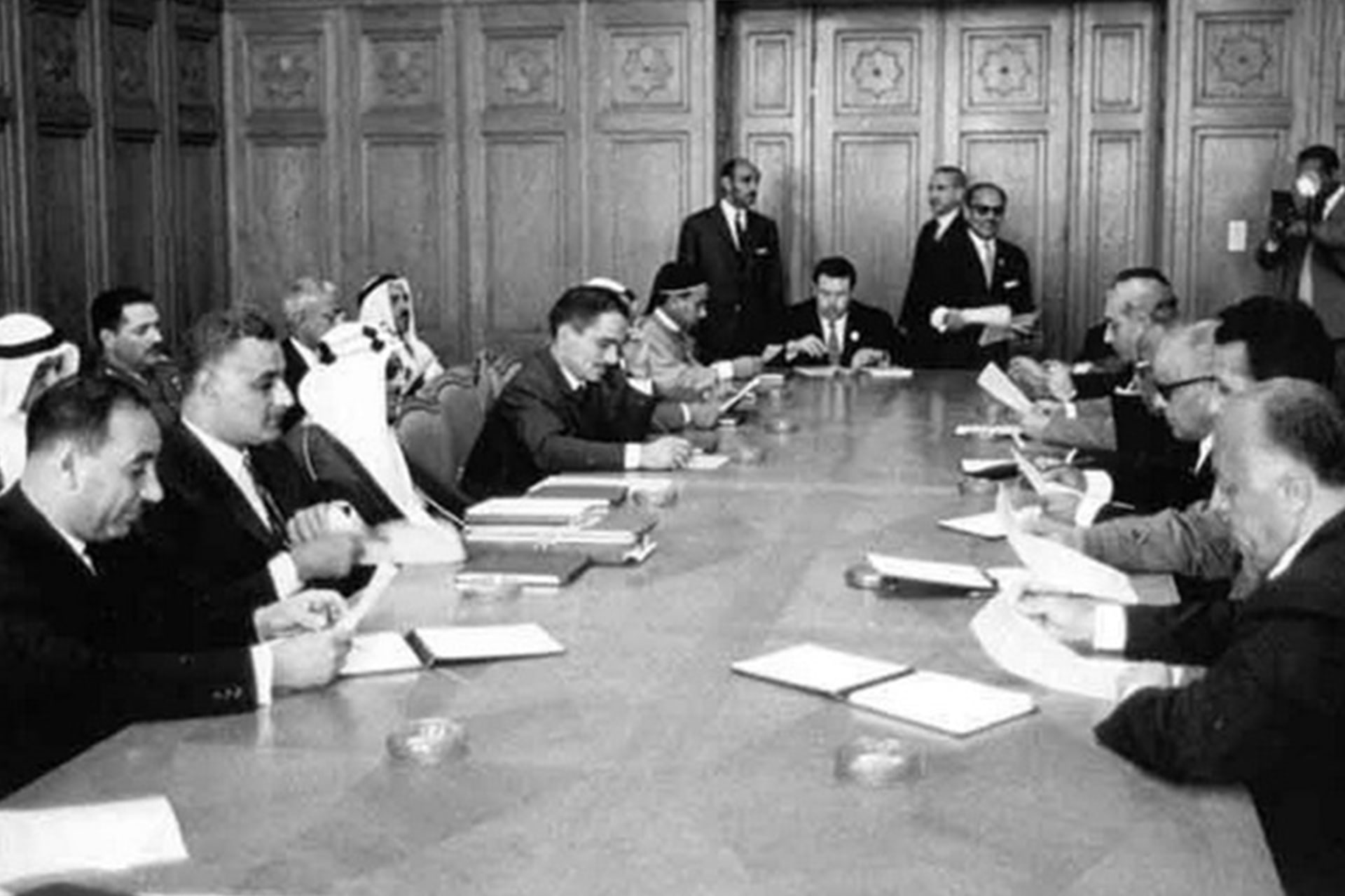 A black and white photo shows men seated at a table with papers in their hands.
