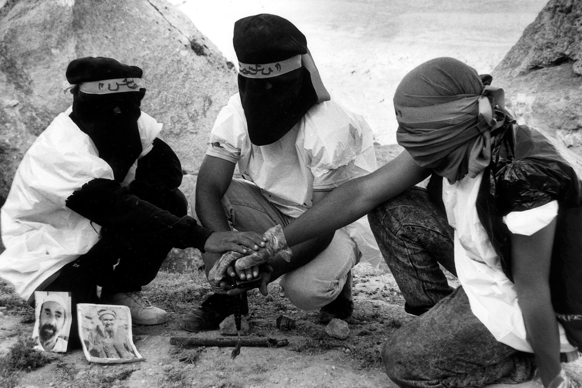 A black and white photo shows a group of three masked figures crouched and stacking their hands on top of one another swearing an oath. Photographs of prominent figures for the movement can be seen next to them.