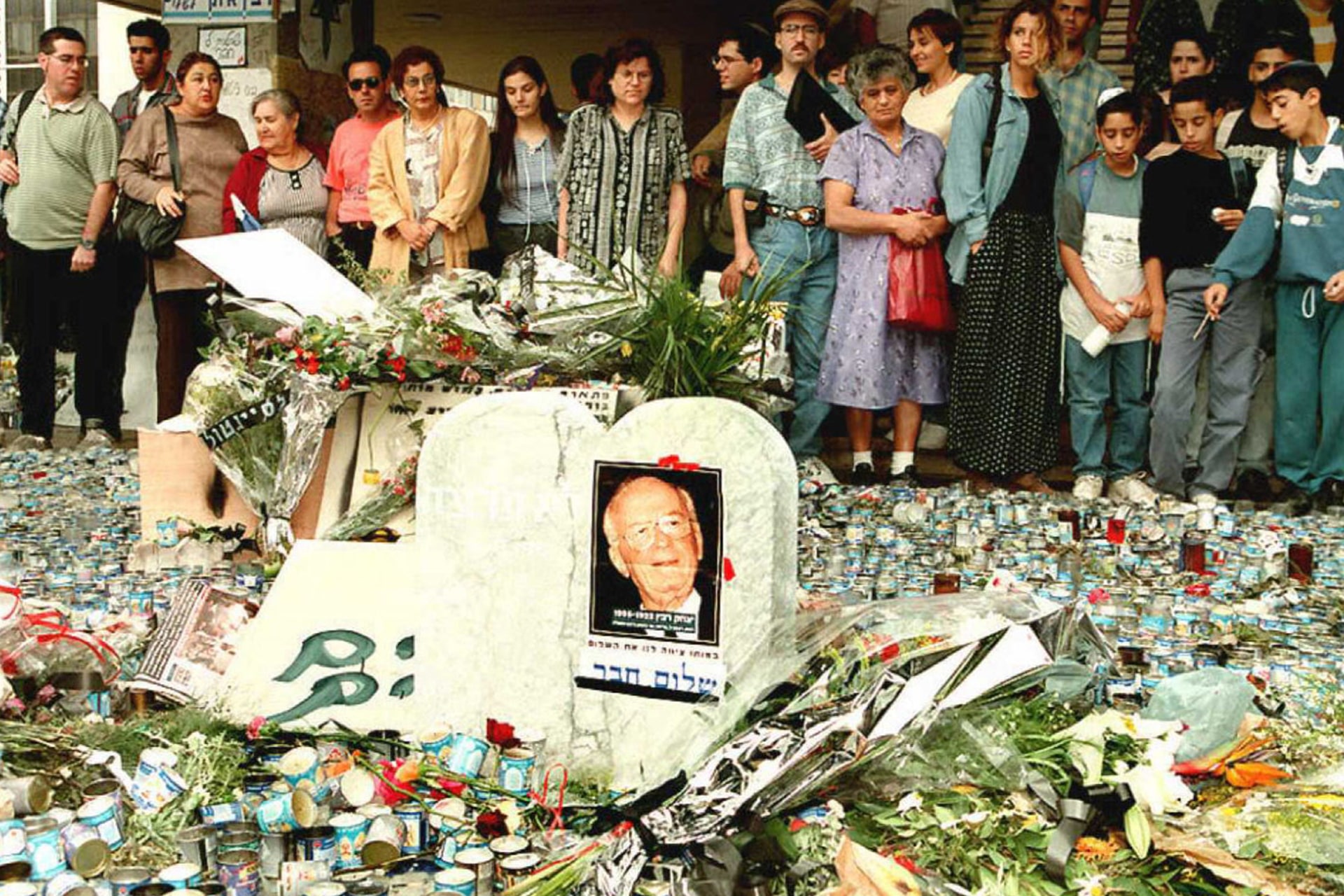A memorial for Yitzhak Rabin can be seen. His photo is placed on a tombstone in the center surrounded by candles, flowers, and signs. People look on solemnly.