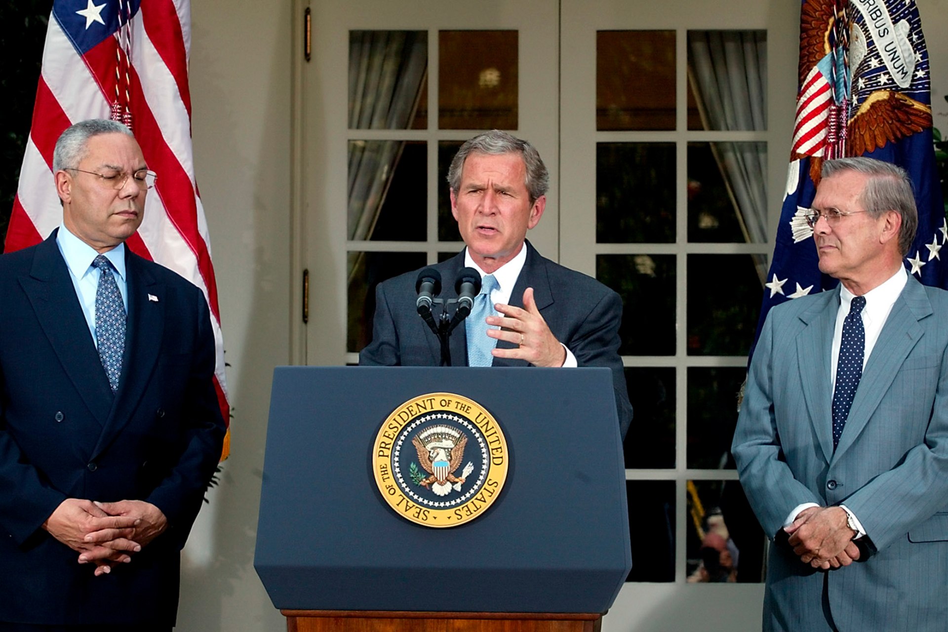 U.S. President George W. Bush (C), joined by U.S. Secretary of State Colin Powell (L) and U.S. Secretary of Defense Donald Rumsfeld (R), speaks at a podium with the presidential seal.