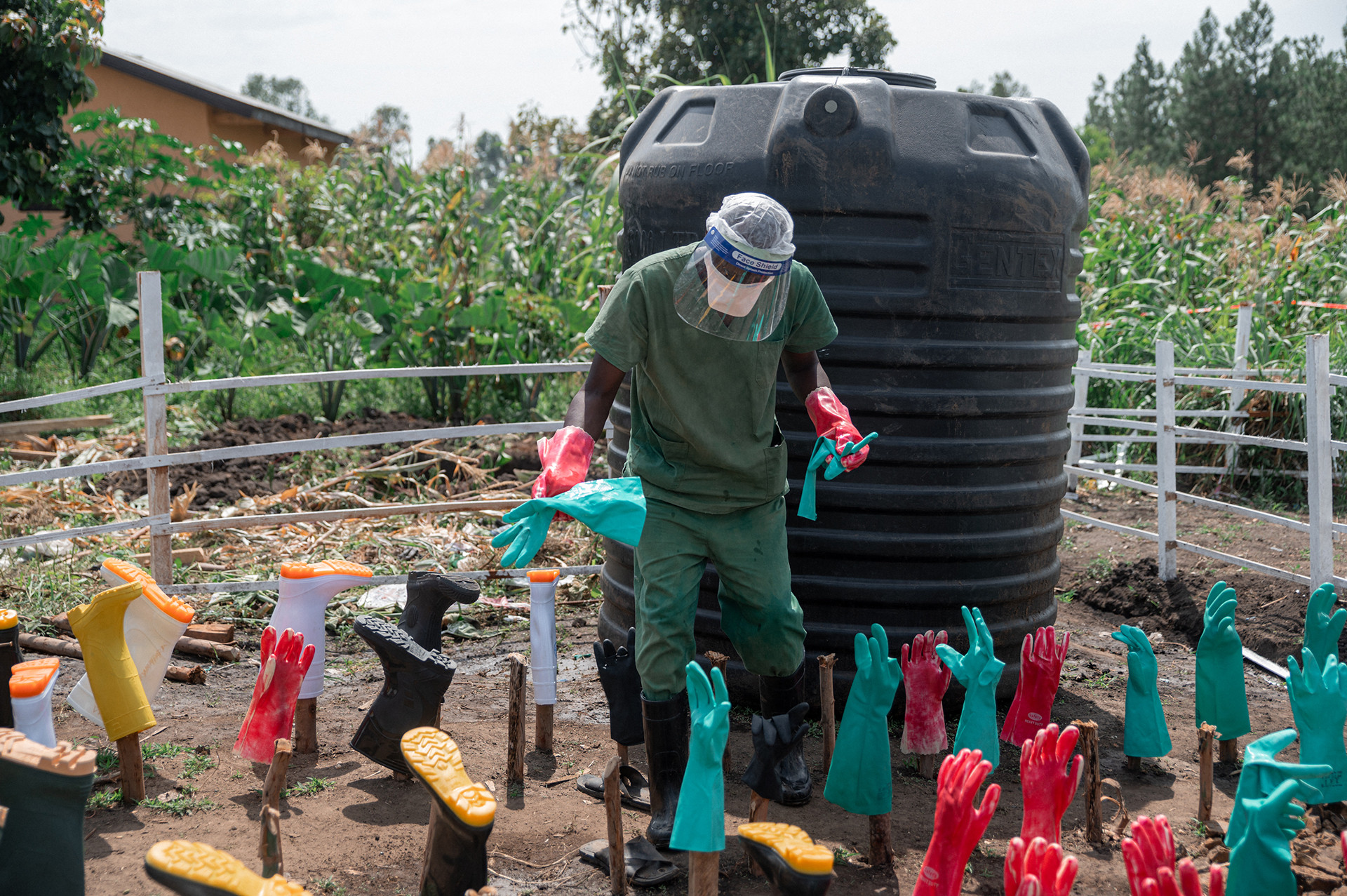 A health worker stands in a fenced-in area in front of a black container, with several stakes in the ground holding up brightly colored rubber gloves and boots that are being disinfected. 