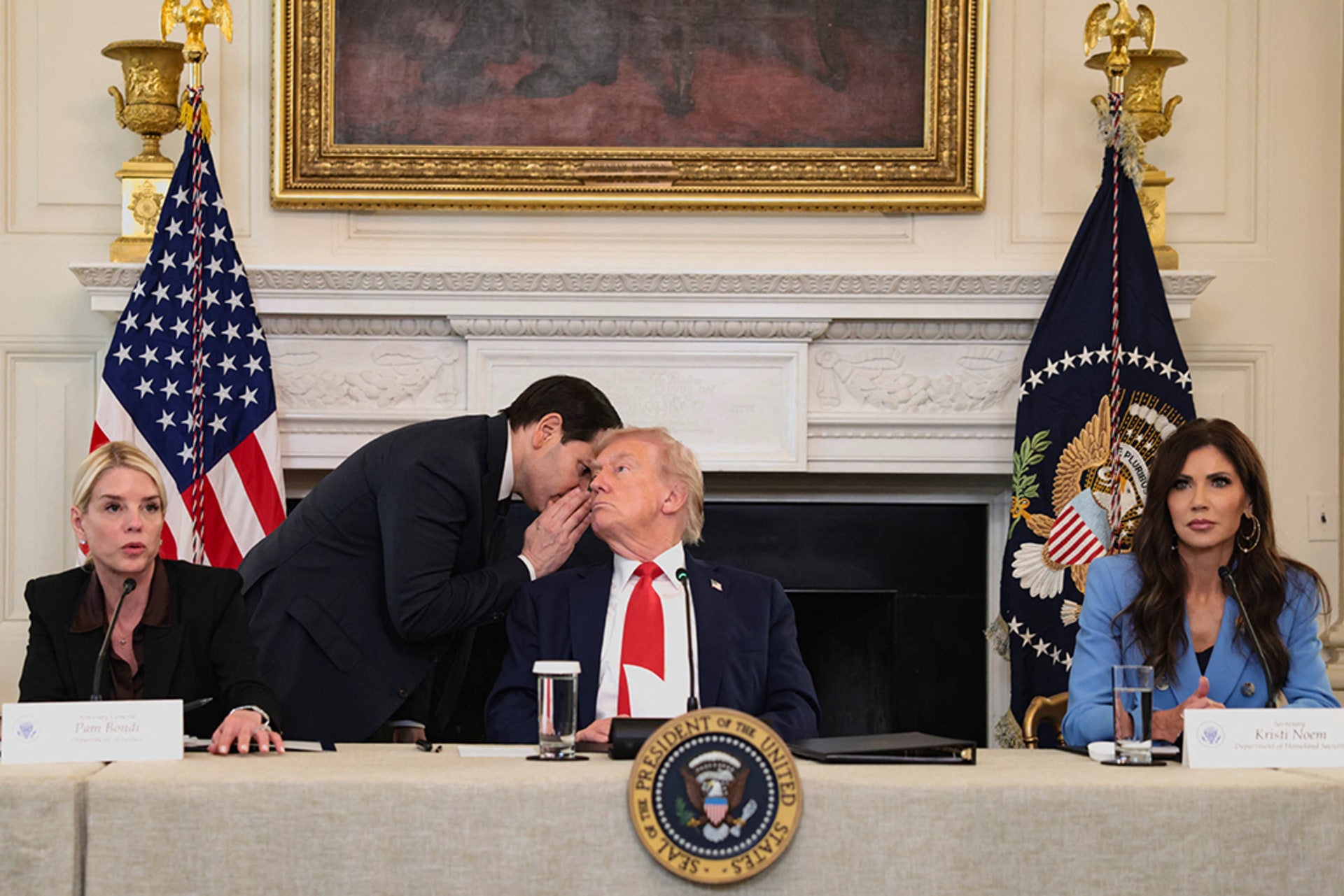 President Donald Trump is seated at a table as Secretary of State Marco Rubio whispers into his ear.