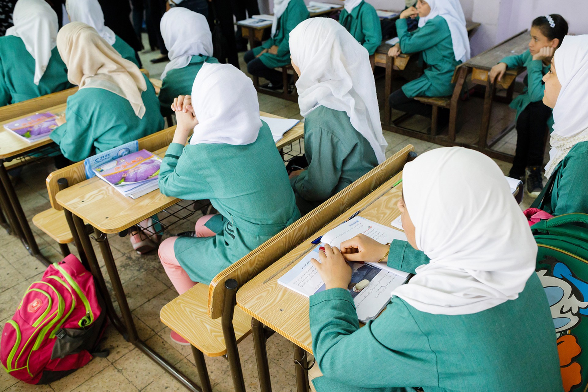 Girls wearing hijabs and school uniforms are sitting on school benches during a lesson in a classroom.