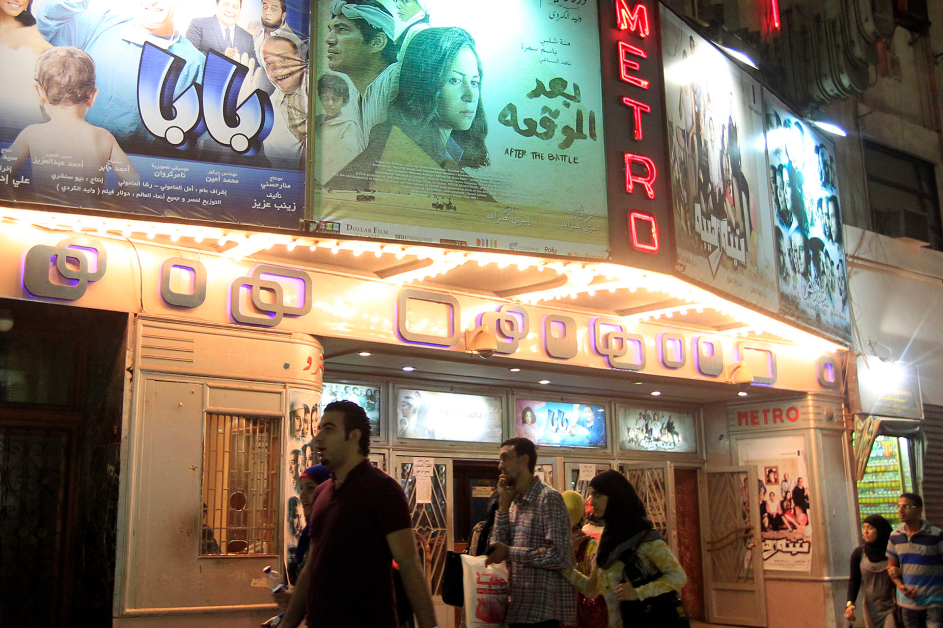 People walk past a cinema called the "Metro" with various posters on the marquee. The marquee is lit up.