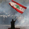 A man waves a Lebanese red and green flag as smoke rises from a street.