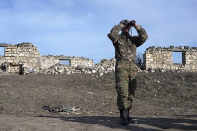 A soldier stands holding binoculars up to his face.