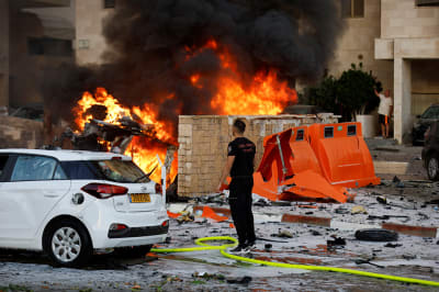 A man stands facing a burning building next to a white car.