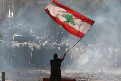 A man waves a Lebanese red and green flag as smoke rises from a street.