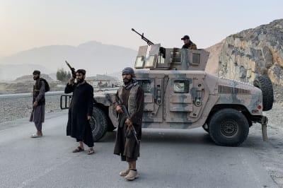 Taliban men holding guns next to a tank with mountains in the background.