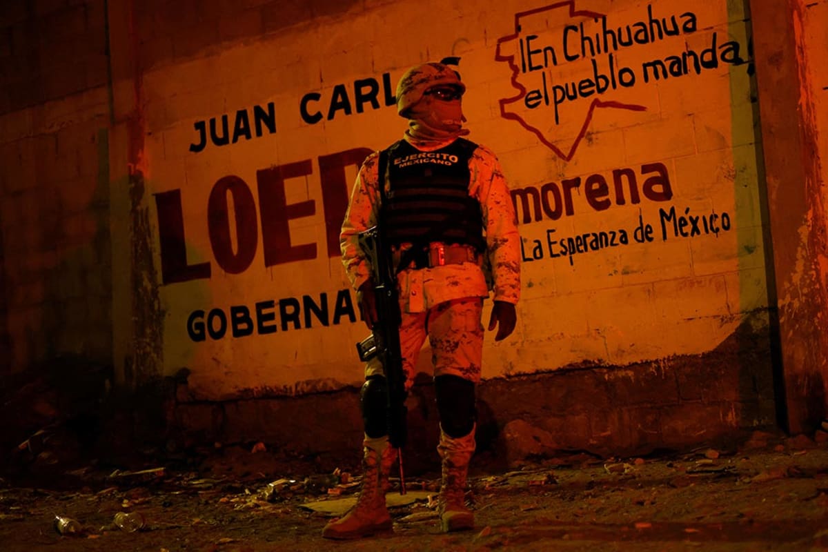 A person in military fatigues stands against a wall facing the camera.