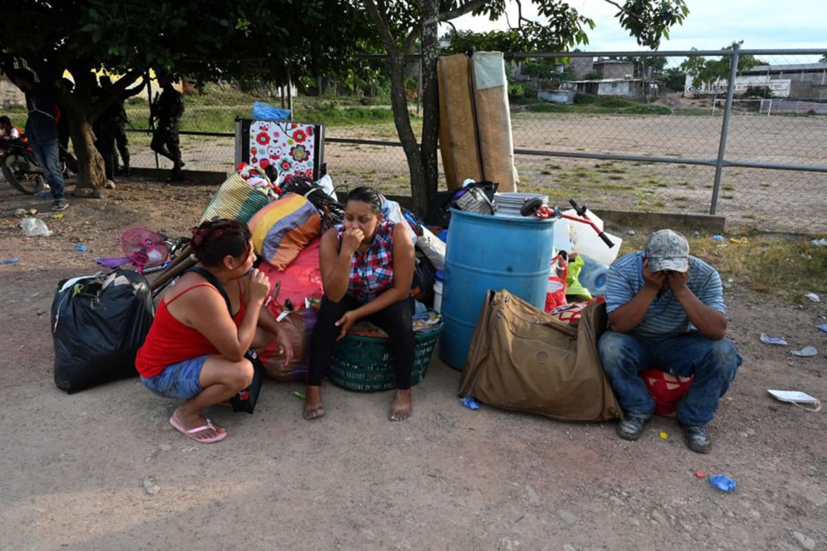 Two women and a man sit on top of their belongings with their hands to their faces.