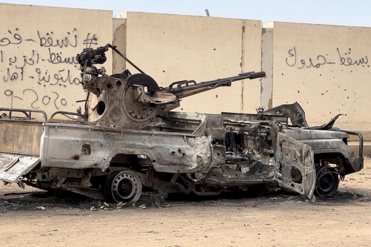 An abandoned, burned-out military tank sits in front of a concrete wall.