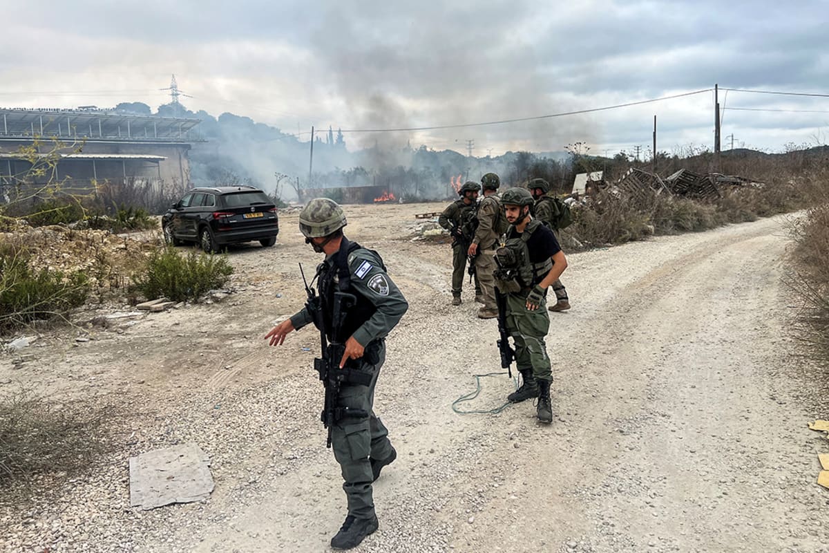 Soldiers stand on a gravel road while smoke billows in the background.