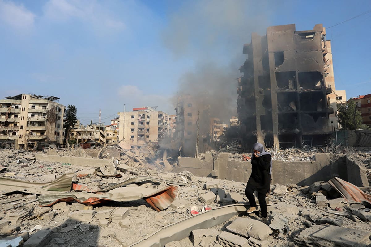 A woman wearing blacks walks on the rubble of bombed buildings while smoke rises.