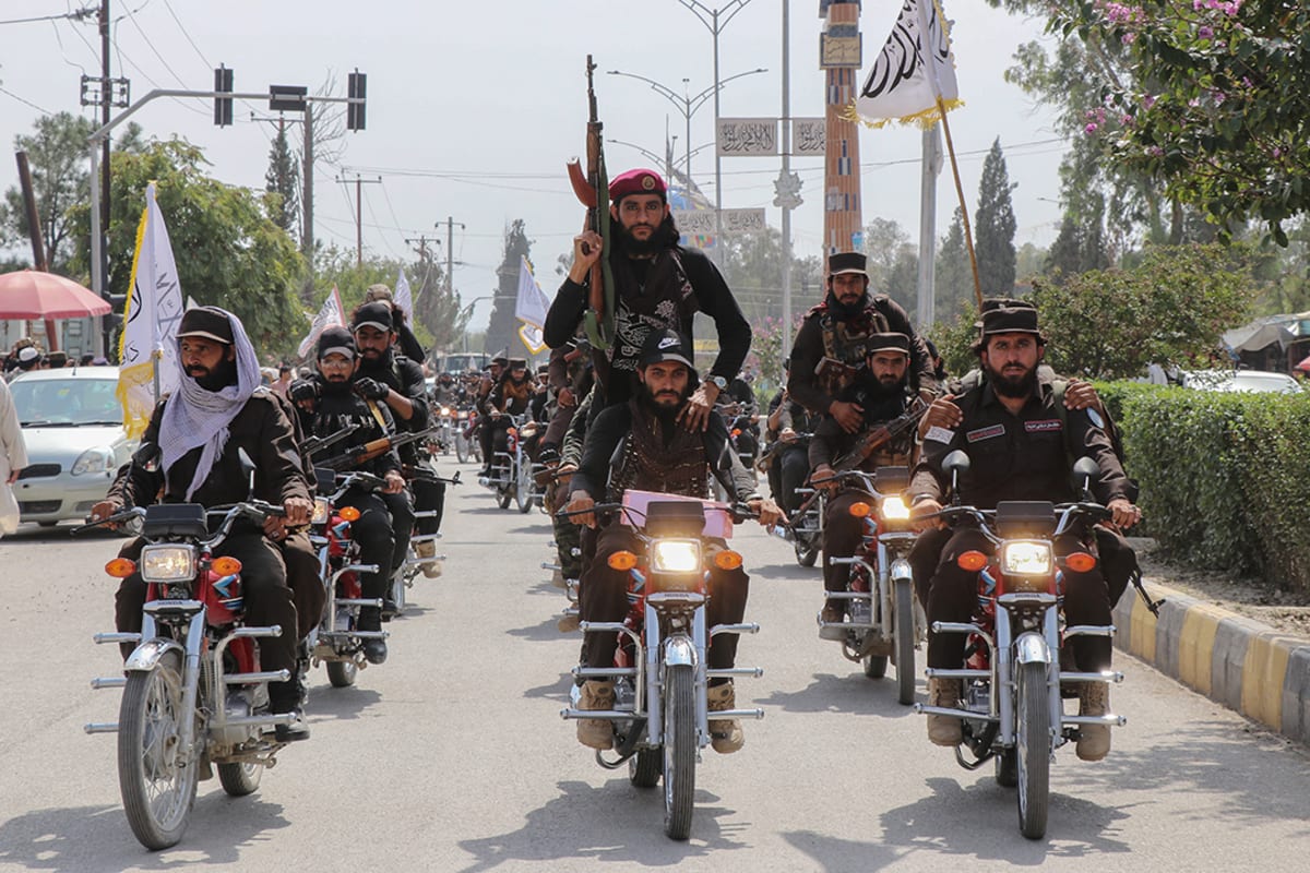 Men ride in formation on motorcycles through an intersection while holding flags and guns.