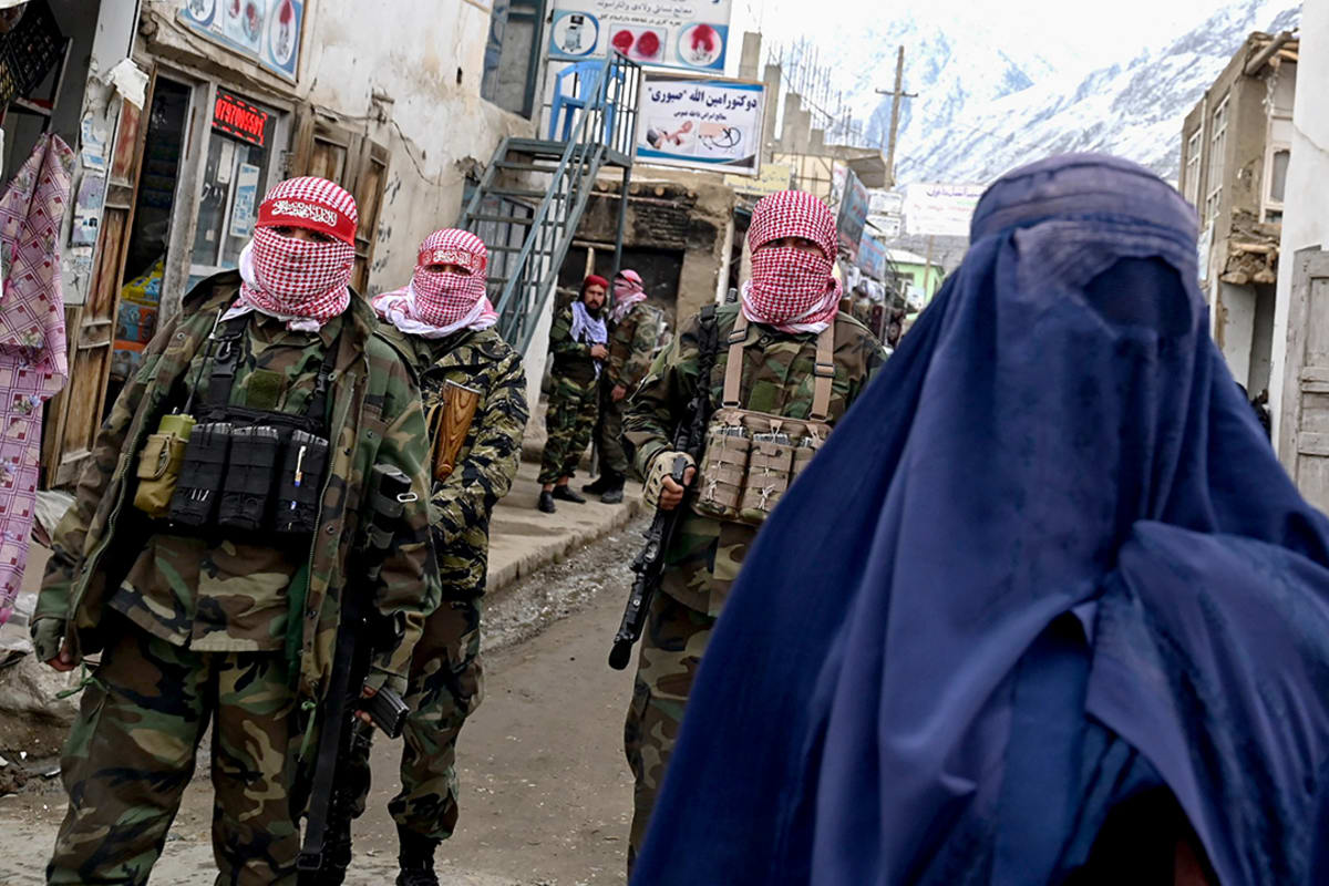 Men in military uniform with stand behind a woman in a blue burqa on a street with mountains in the background.