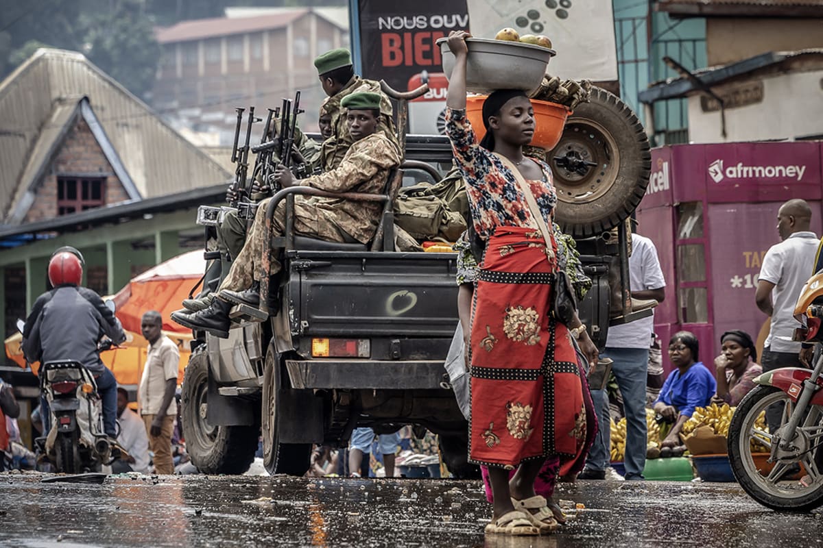 Soldiers sit in a pick up truck while a woman carrying fruit above her head walks by.