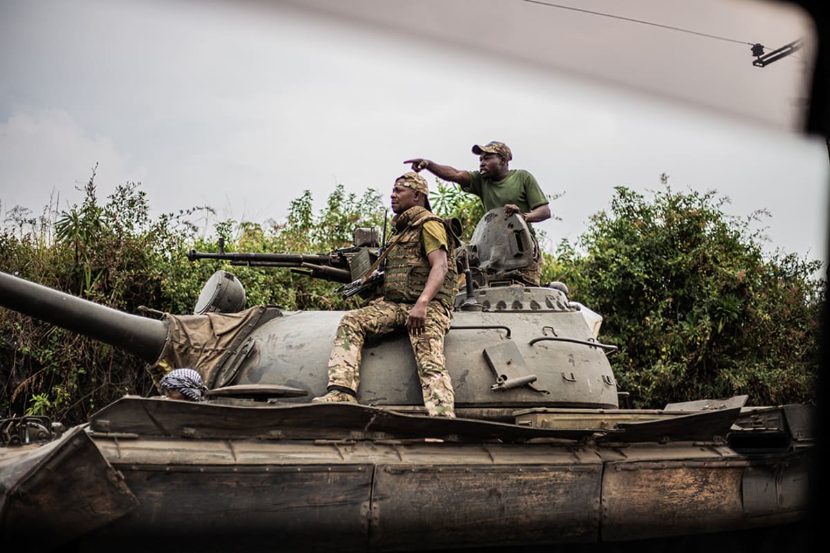 Two soldiers sit on top of a tank while pointing at something ahead of them.